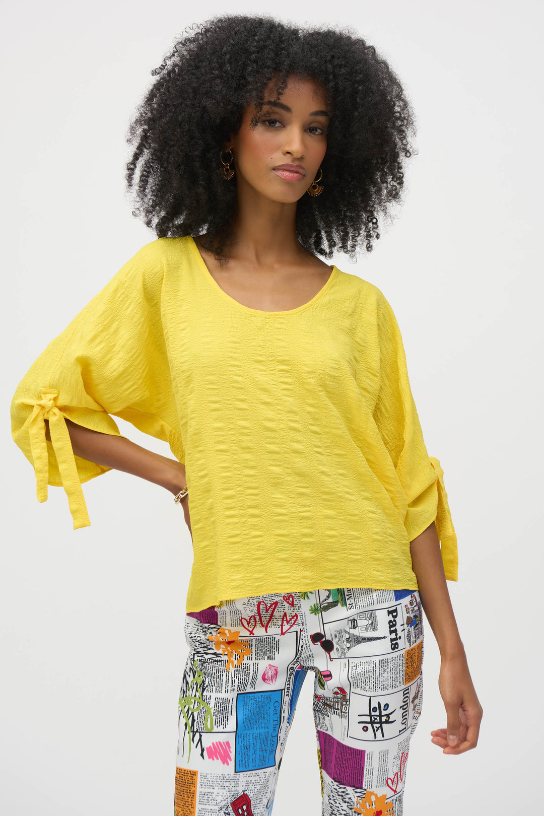 Joseph Ribkoff Spring 2025 Expertly crafted with textured knit fabric, this top offers a looser fit and puffy half sleeves with tie details that make it a standout piece. The scoop neck adds a touch of elegance to this versatile top, perfect for any occasion. 