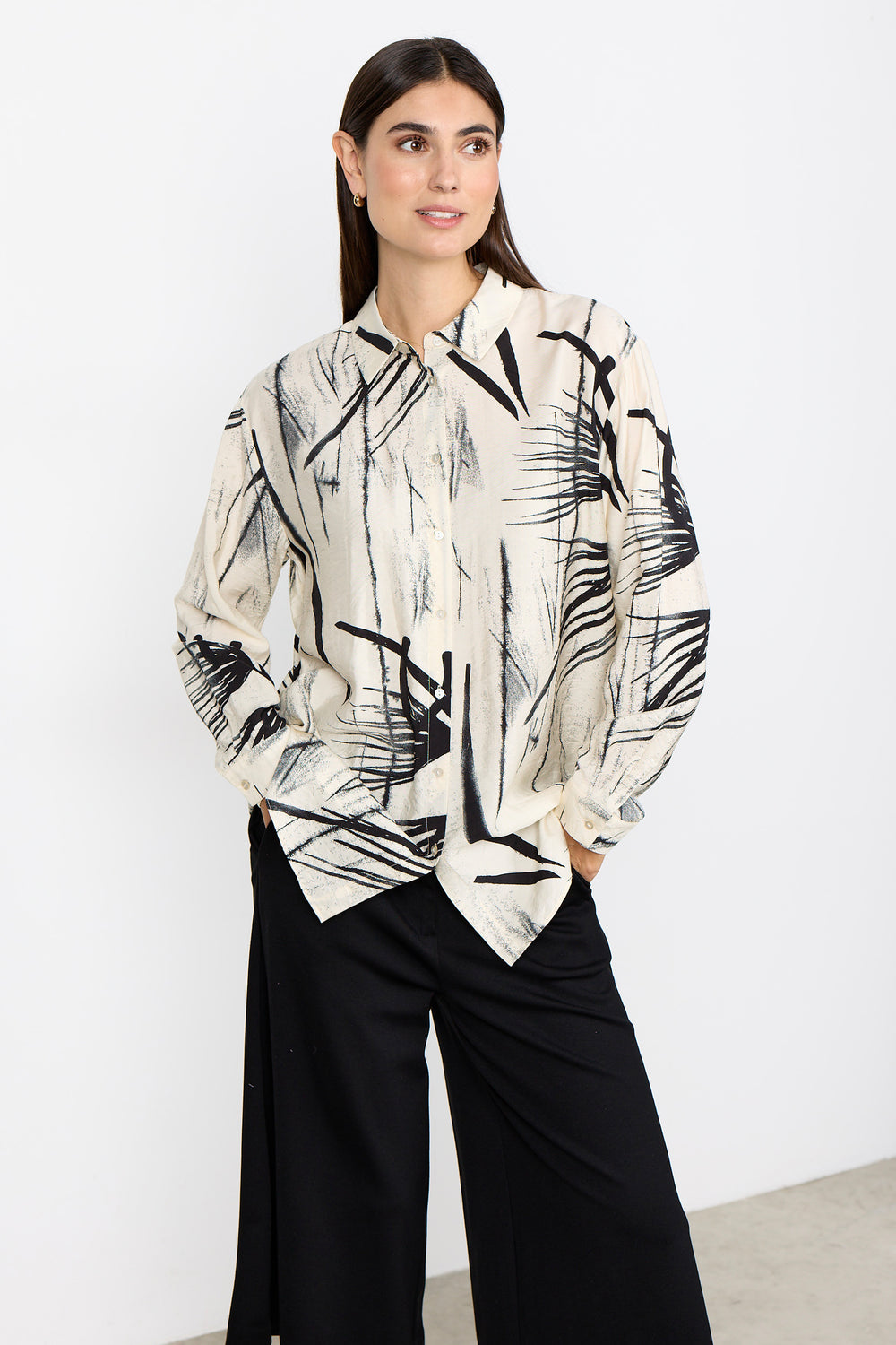 Woman wearing a cream blouse with abstract print and black pants on a white background. 